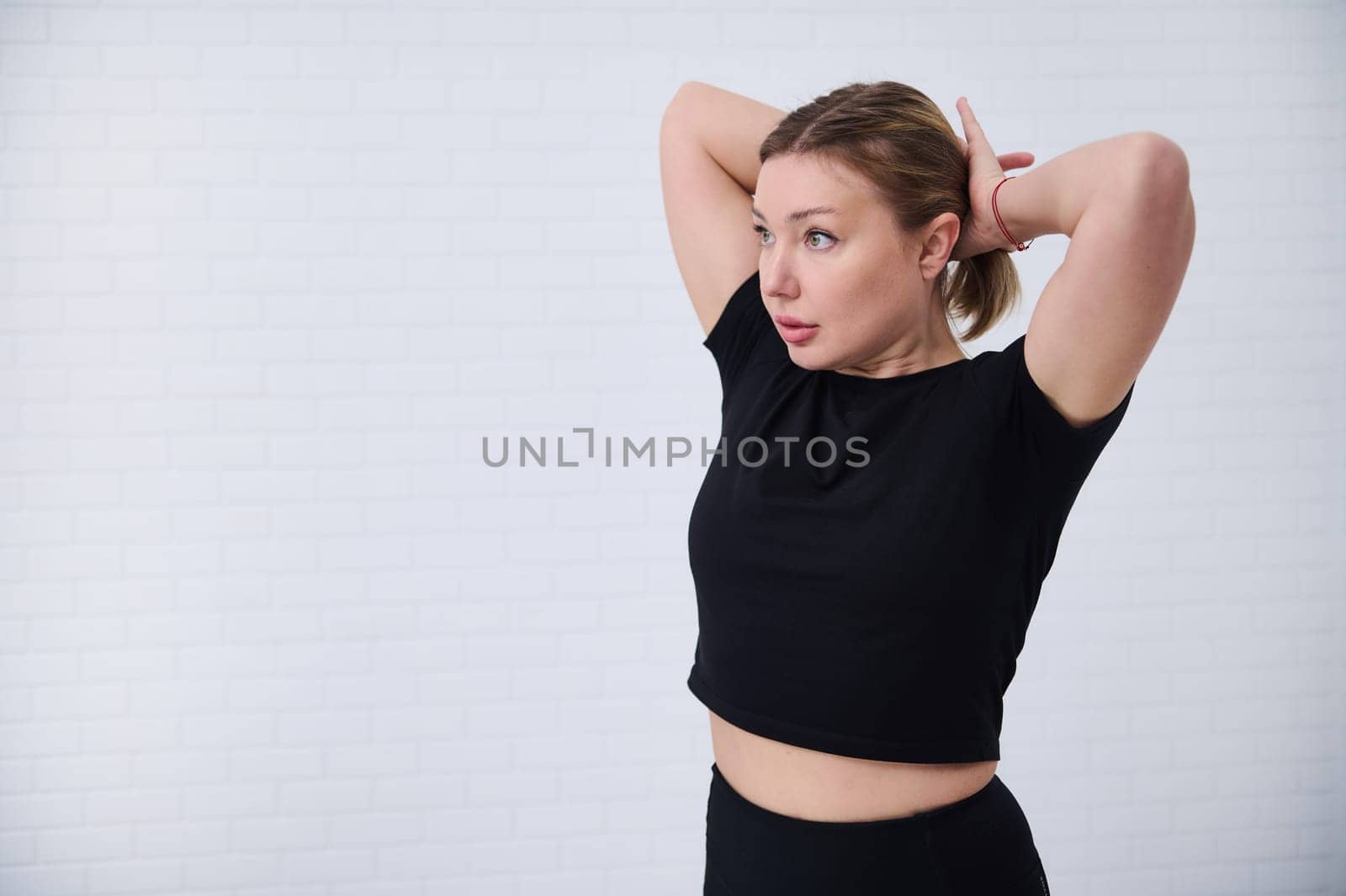 Beautiful Caucasian European athletic woman 30s in black sportswear, tying a ponytail, isolated over white wall background