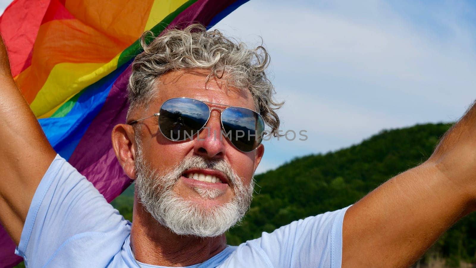 Portrait of a gray-haired elderly Caucasian man with a beard and sunglasses holding a rainbow LGBTQIA flag against a sky background, shouts in protest, Celebrates Pride Month Coming Out Day