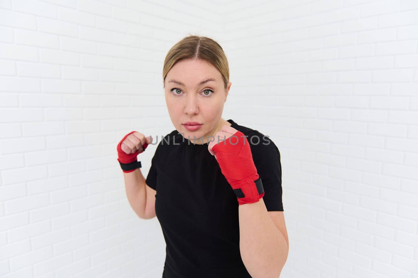 Confident waist up shot of a strong young woman punching forward a camera with boxing gloves, practicing martial art, box, kickbox isolated on white background. Sport. Cardio workout. Endurance.