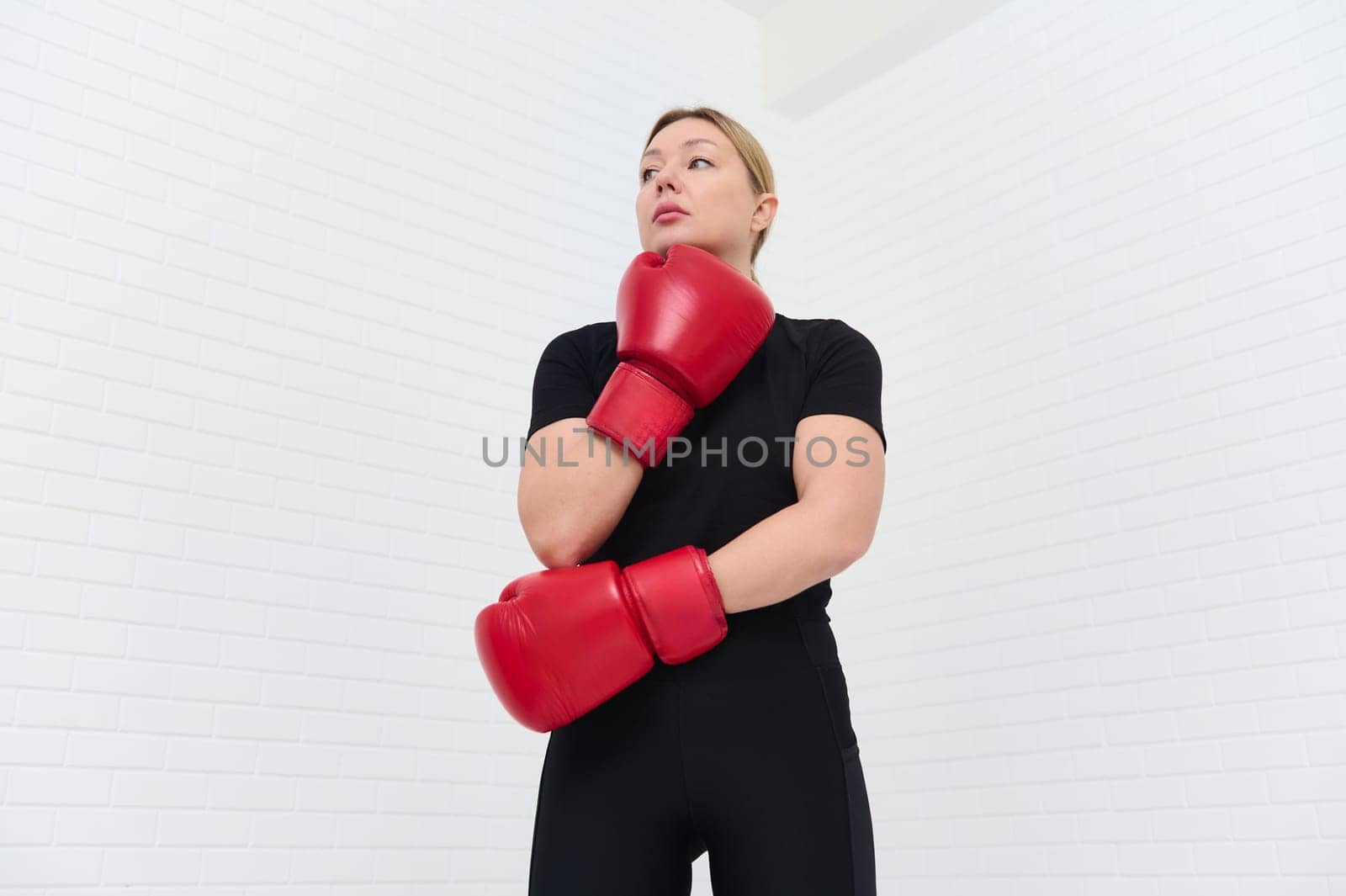 European confident woman boxer with red gloves and black sports wear, posing against white background. View from the bottom. Concept of female strength, combat training, and athletic discipline