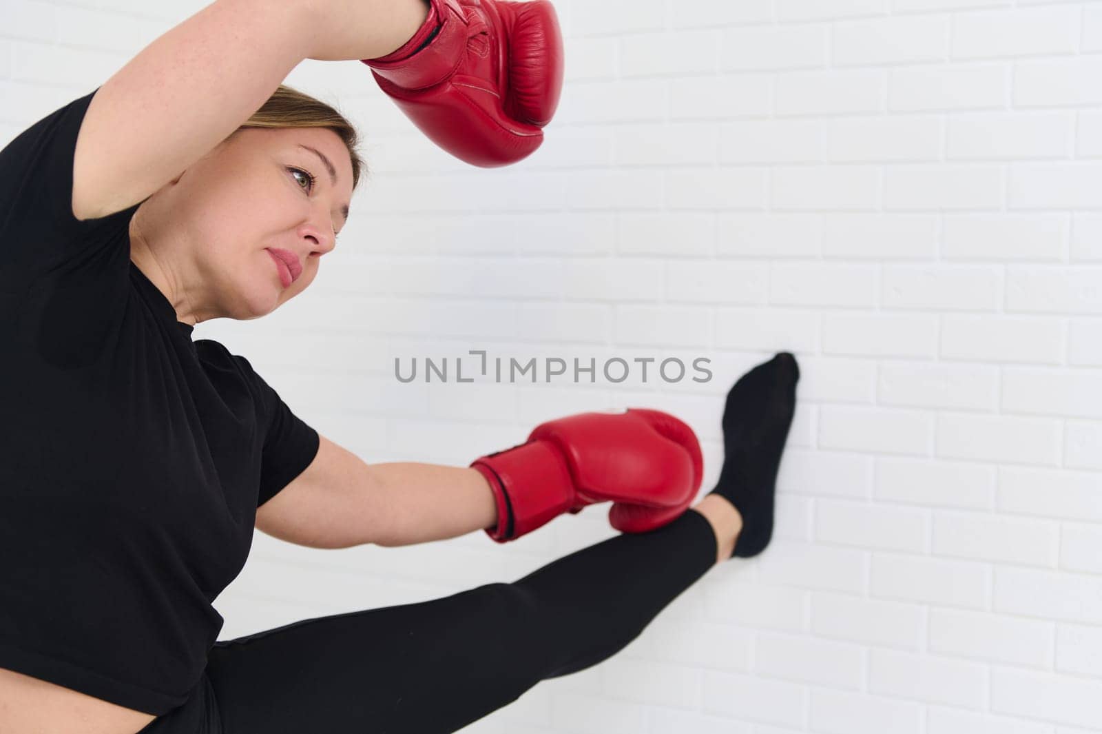 Female boxer stretching arms, warming up her body before intensive sport, wearing boxing gloves over white studio background. Sport. Cardo workout. People and active healthy lifestyle concept