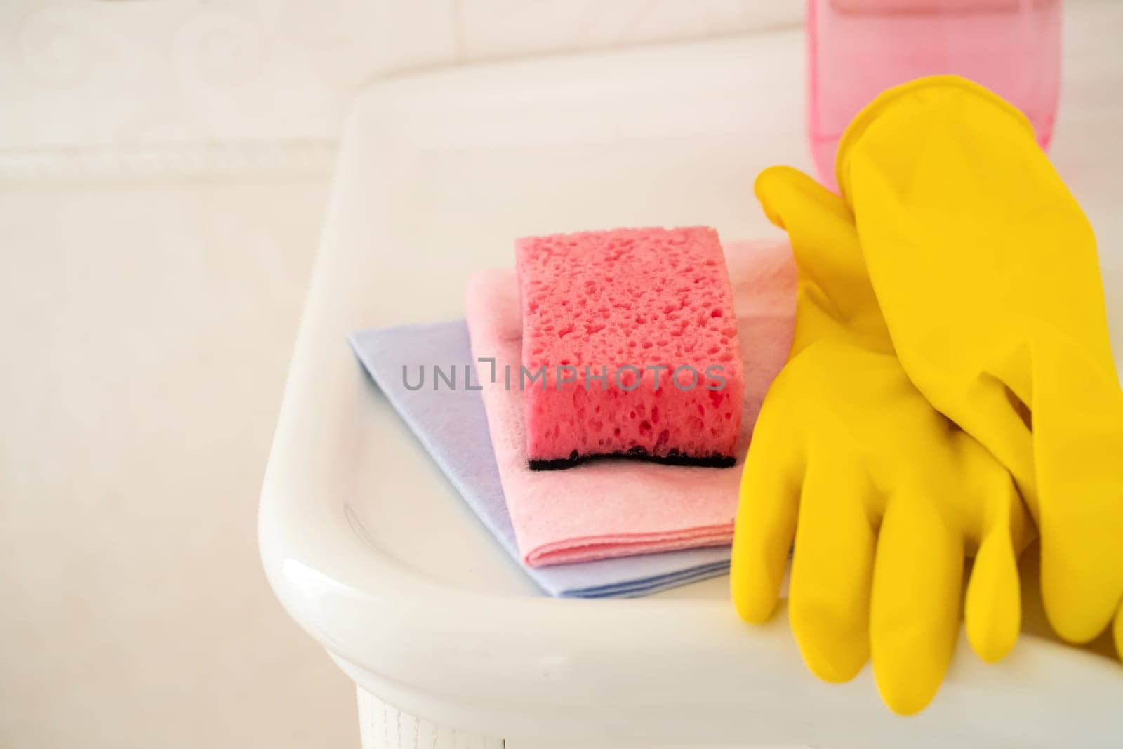 Household cleaning items, such as rubber yellow gloves, several types of rags and sponges, lie on a clean white washstand in the bathroom.