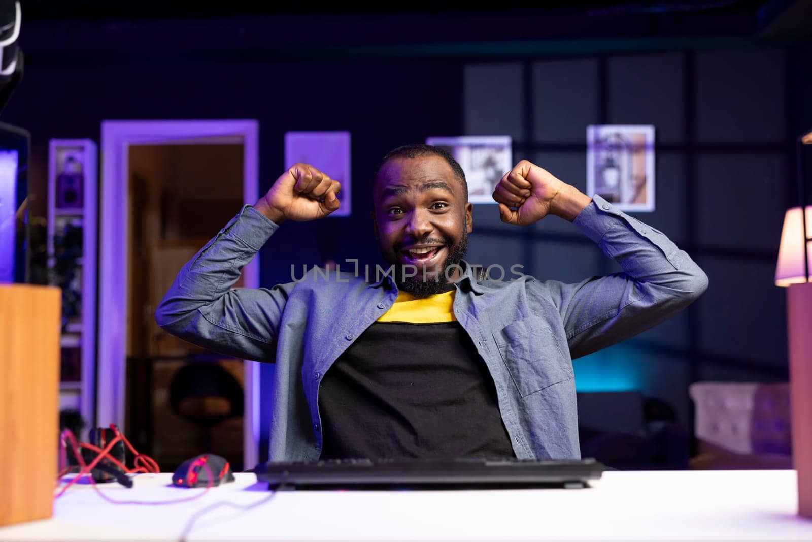 Portrait of delighted BIPOC man on desk chair celebrating winning videogame level in apartment. Happy african american player celebrating after achieving victory in game on gaming PC