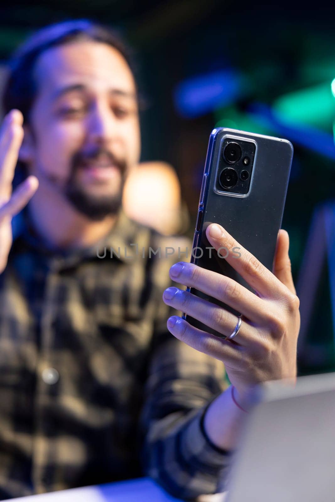 Male individual waving towards cell phone, making video call with colleagues. Closeup of youthful man sitting and having an online conference call with clients on mobile device.