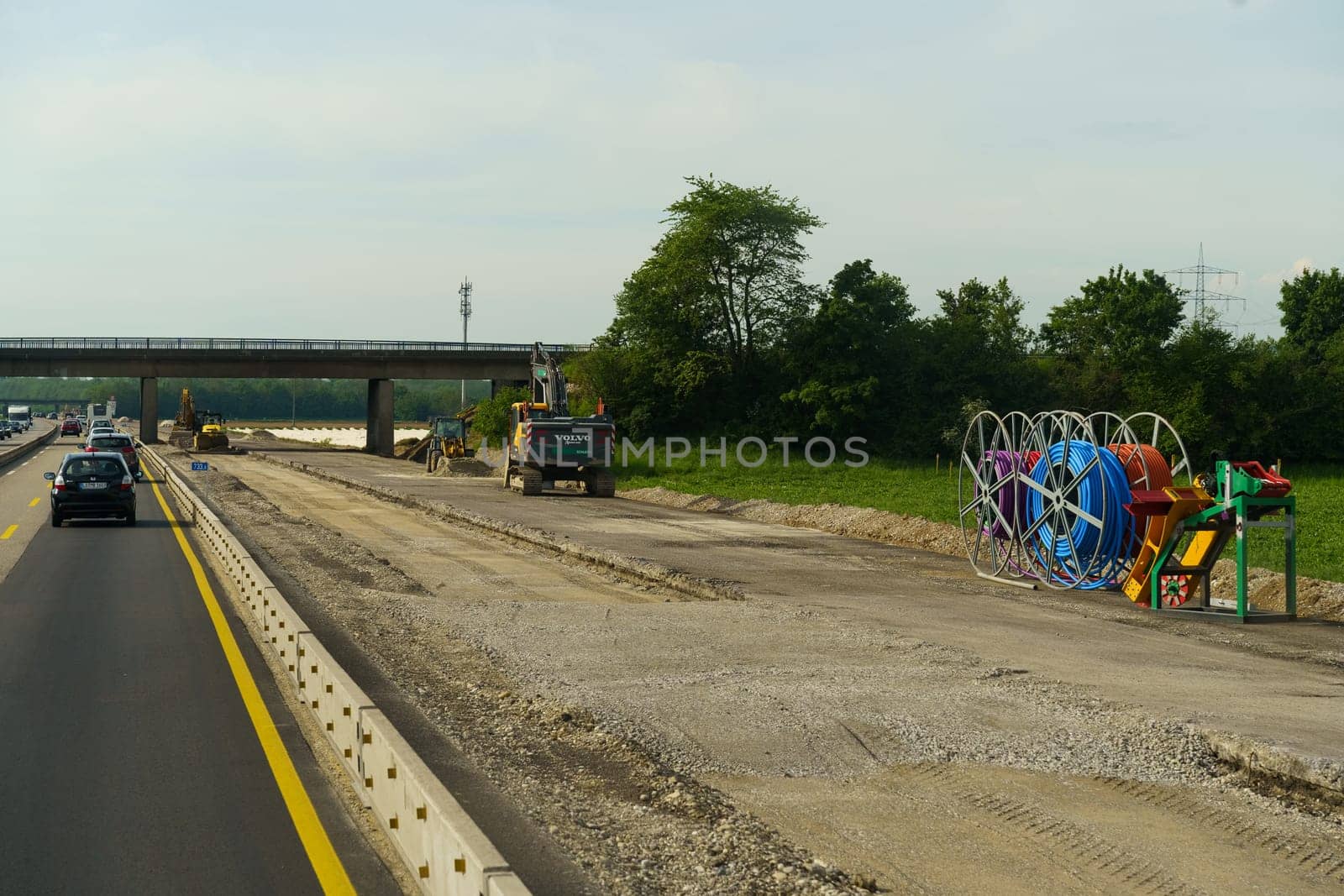 Weimar, Germany - May 7, 2023: Highway construction workers are seen using machinery while vehicles pass by. The focus is on road construction work near a scenic green area with an overpass in the background.