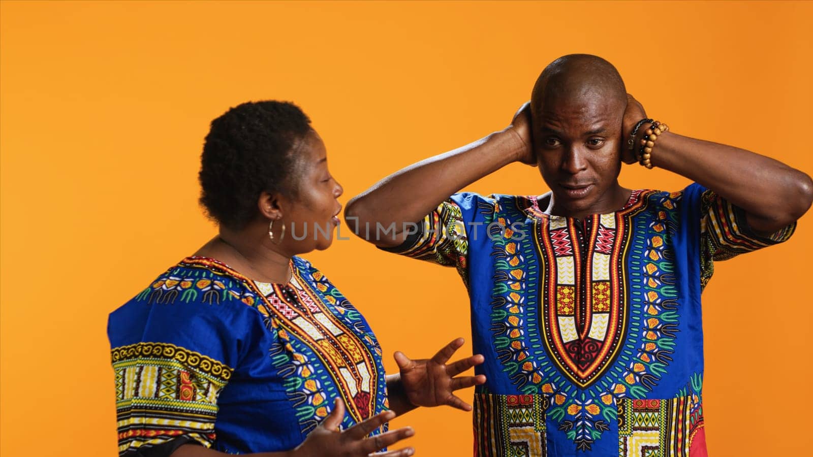 Furious girlfriend shouting at her boyfriend and fighting, having disagreement over orange background. Frustrated man covering his ears and trying to ignore his wife yelling in studio.