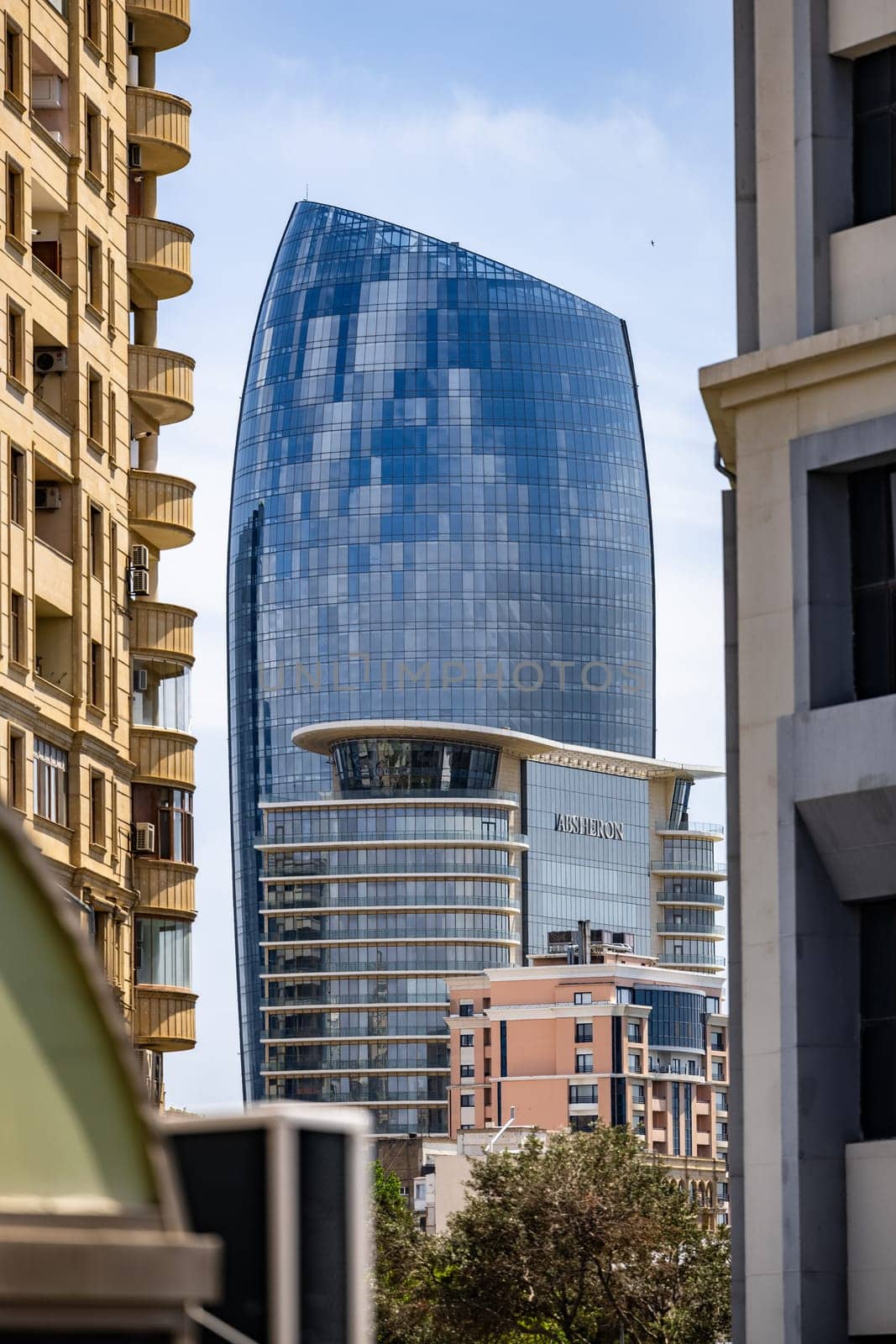 A tall building with a glass facade is seen from the street. The building is surrounded by other buildings, and there are trees in the background. The sky is clear and blue