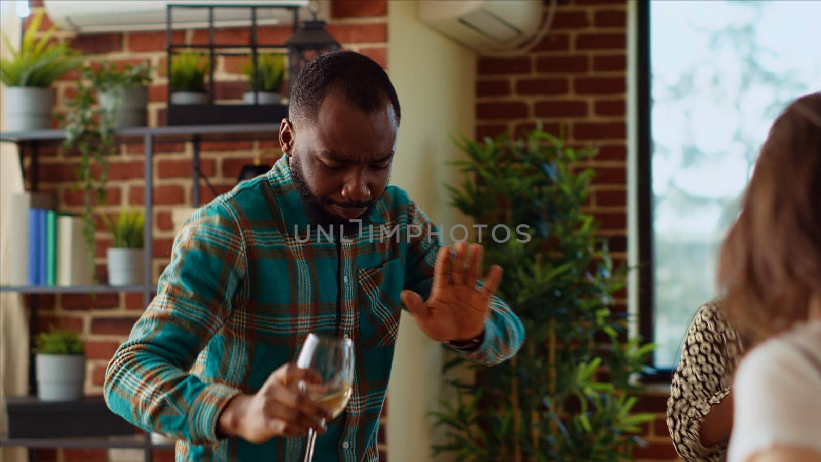 Guests at home party doing dance moves in brick wall living room, holding wine glasses in hand. African american man dancing with friends on energetic beats during weekend hangout, shaking hips