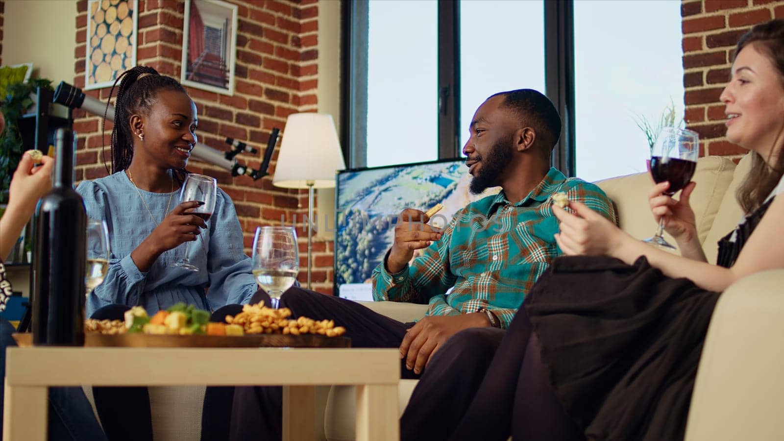 Interracial couple at social gathering talking on couch while enjoying glass of wine. Husband and wife guests at apartment party having pleasant conversation in brick wall home, eating snacks