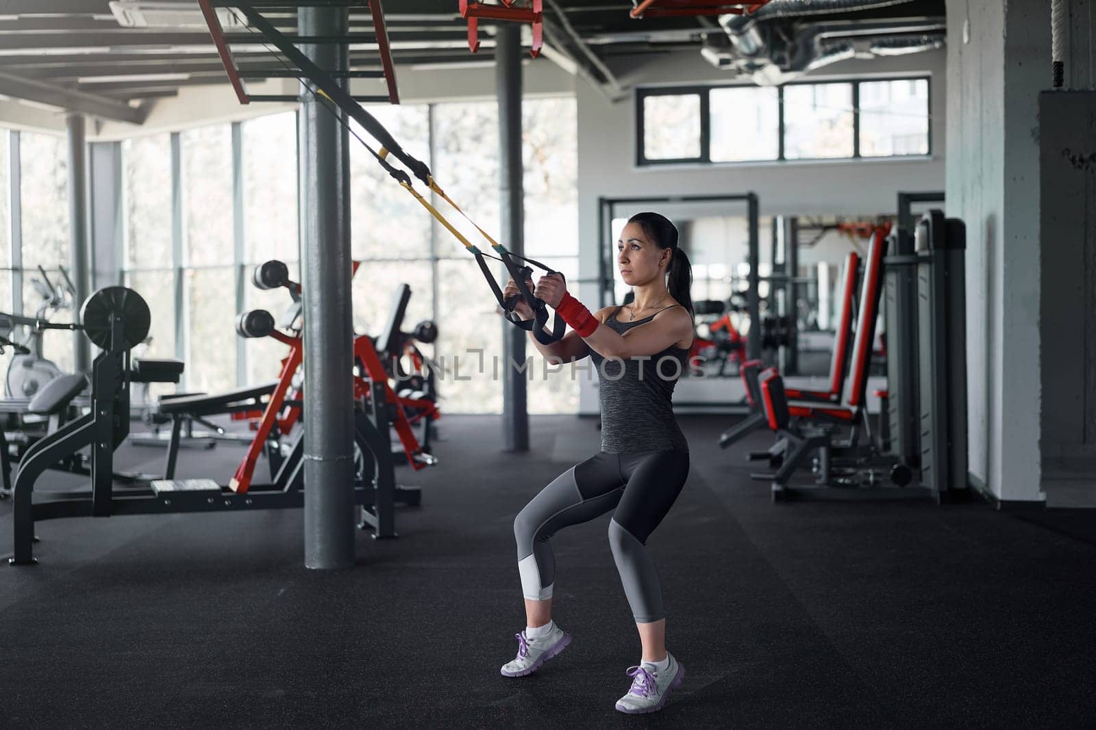 Full body of athlete woman training indoors with suspension system, doing stretching and strength exercises using special straps. Caucasian sportswoman using resistance bands in gym.
