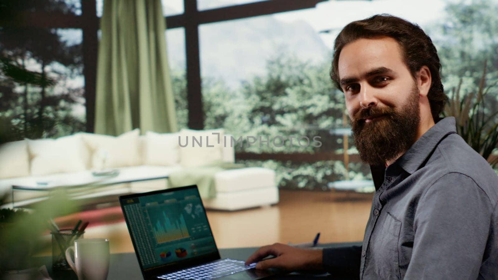 Young man sitting at desk in his luxury cabin with large windows showing panoramic forest view and opening pc, working on laptop thinking about new income development. Billionaire at mountain villa.