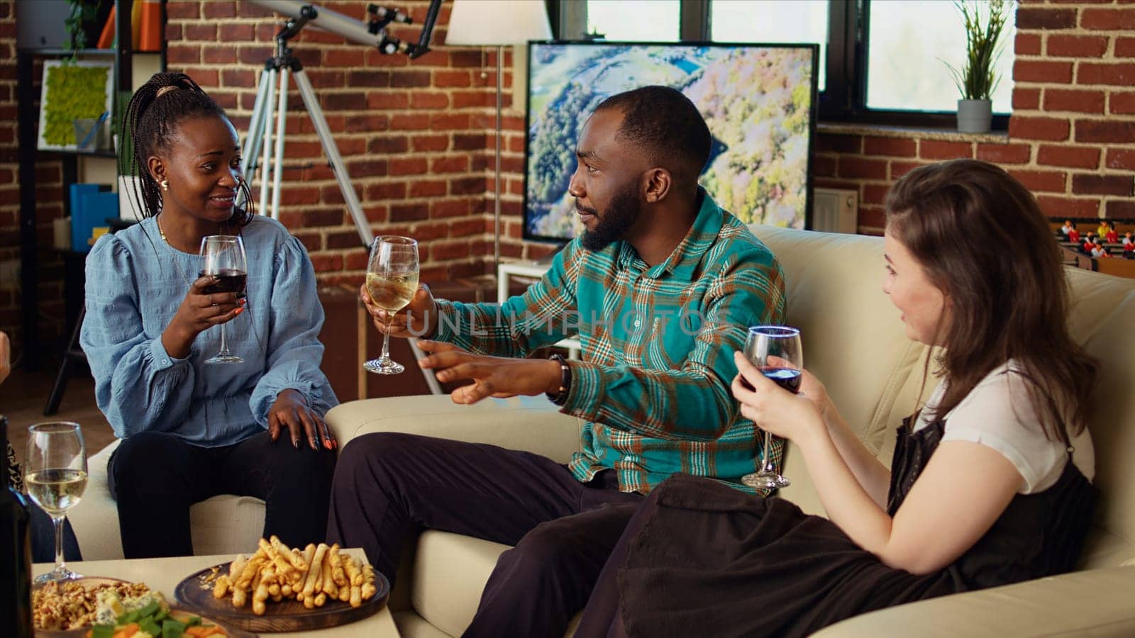 Guest making multiracial people laugh in cozy house while drinking alcohol and eating snacks by DCStudio