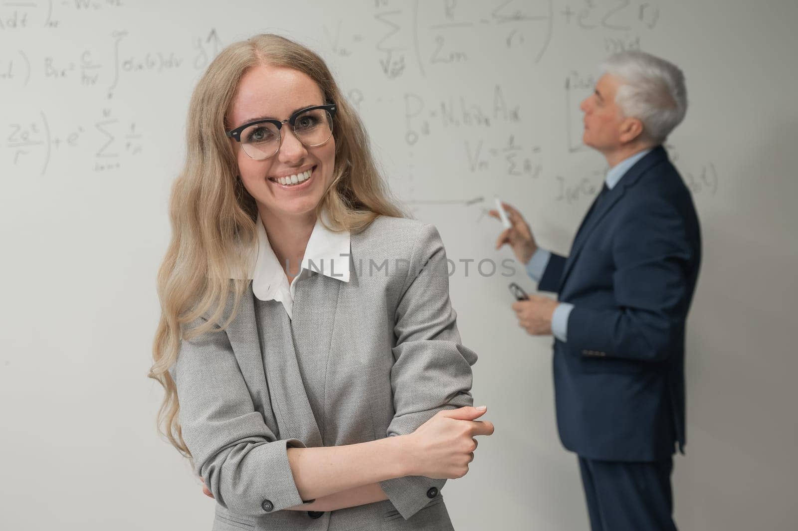 An elderly man writes on a white board and a young woman stands thoughtfully