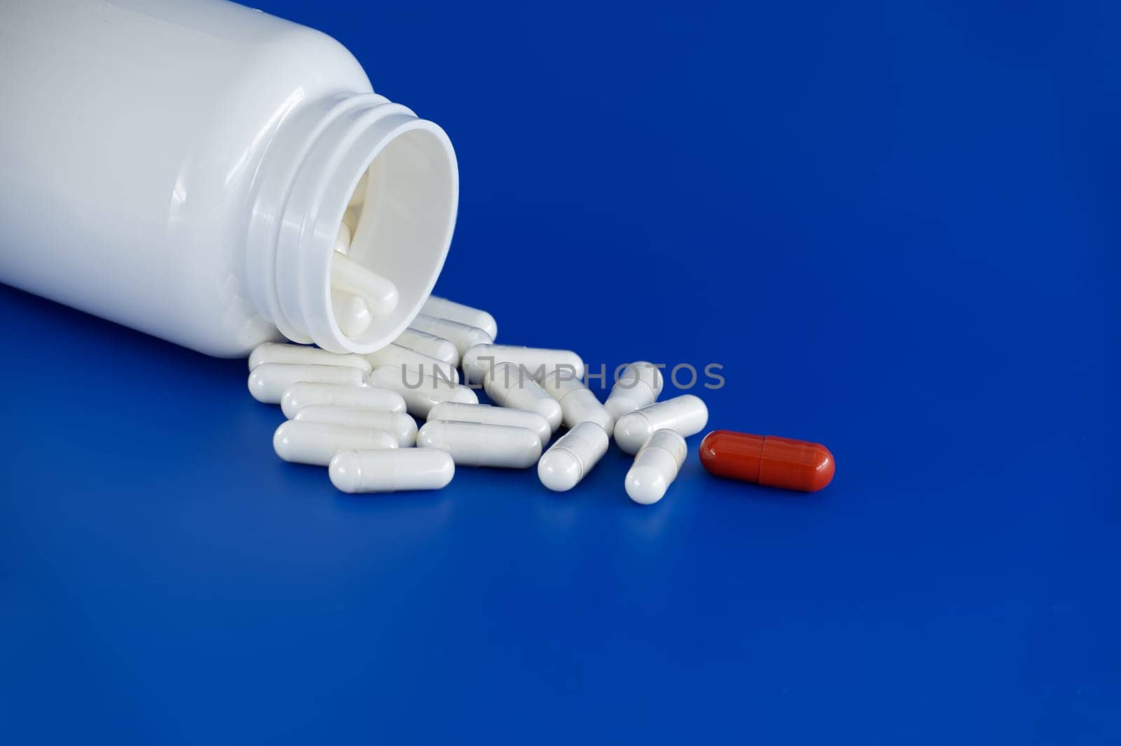 Overturned white medicine bottle with its contents white pills or capsules, spilled out onto a blue background. Among these white pills, a solitary red pill stands out