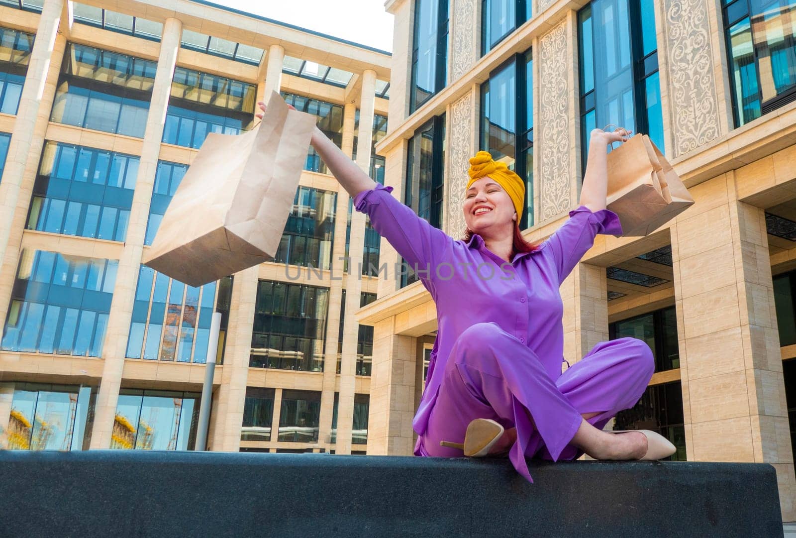 woman with craft shopping bags in the city. The concept of fashionable Consumerism and ecology. An adult Asian woman in a purple suit with red and yellow accents enjoys shopping . a sunny day.