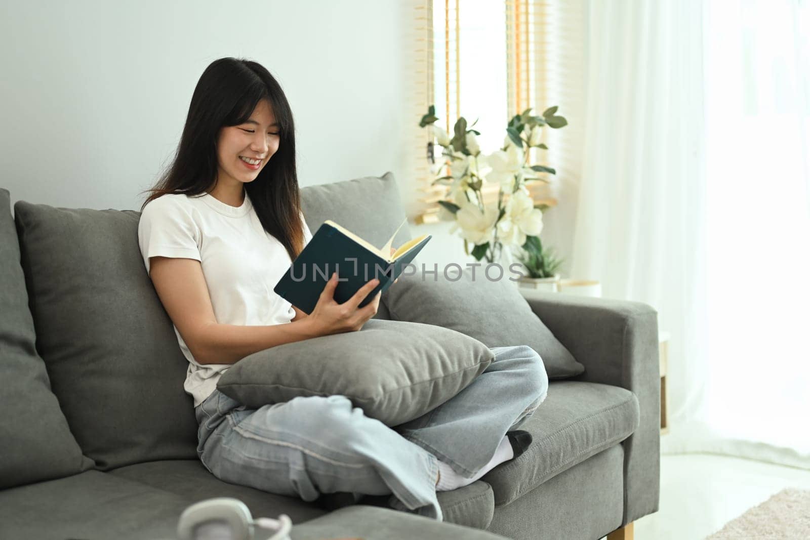 Smiling young woman sitting on cozy sofa and reading book. People, leisure and lifestyle concept.