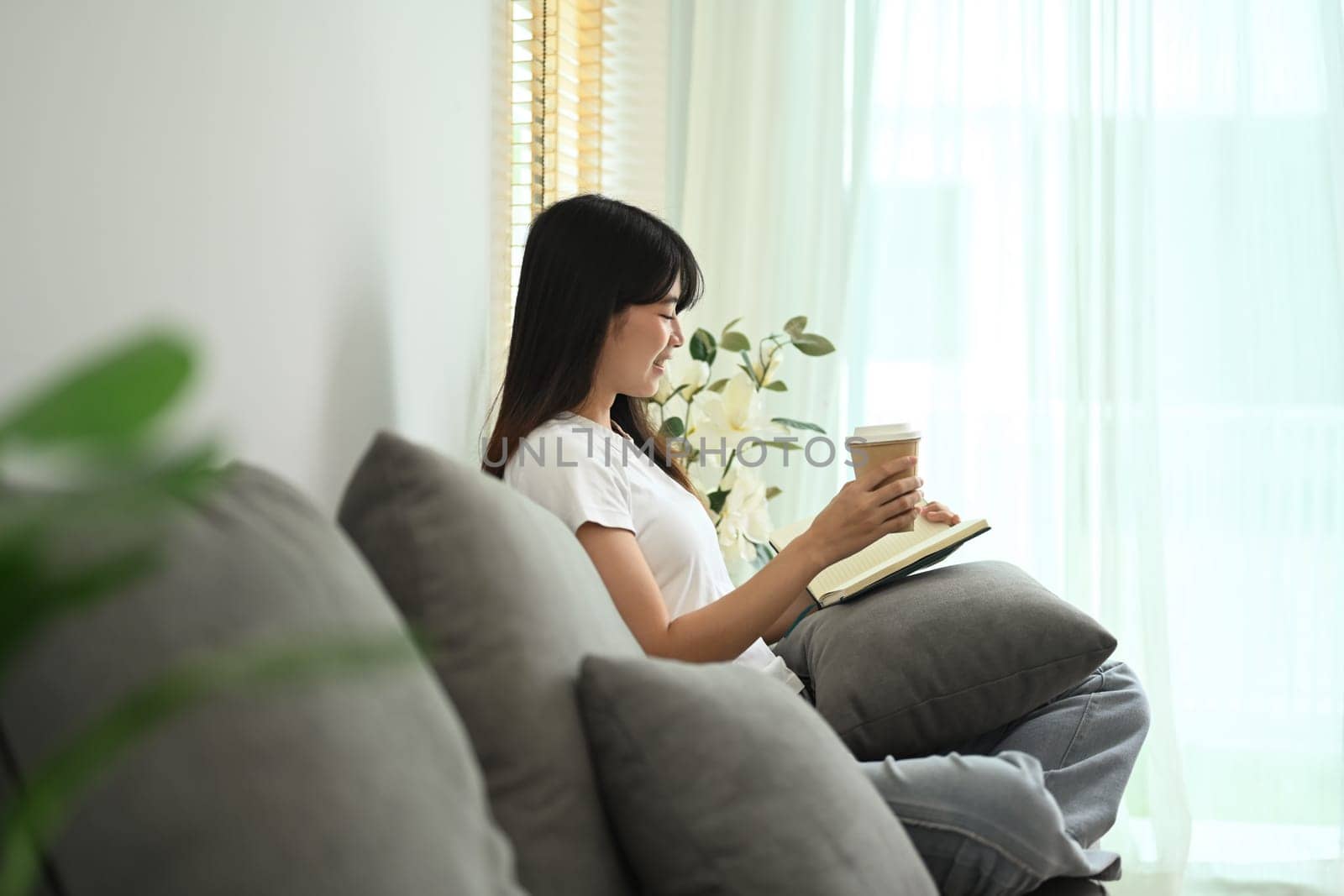 Peaceful young woman with paper cup of coffee reading book on couch at home.