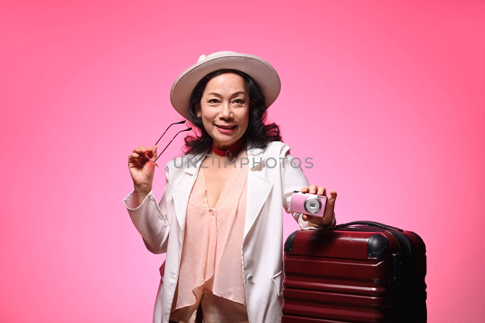 Cheerful senior woman with travel accessories on pink background. Summer, travel and vacation concept.