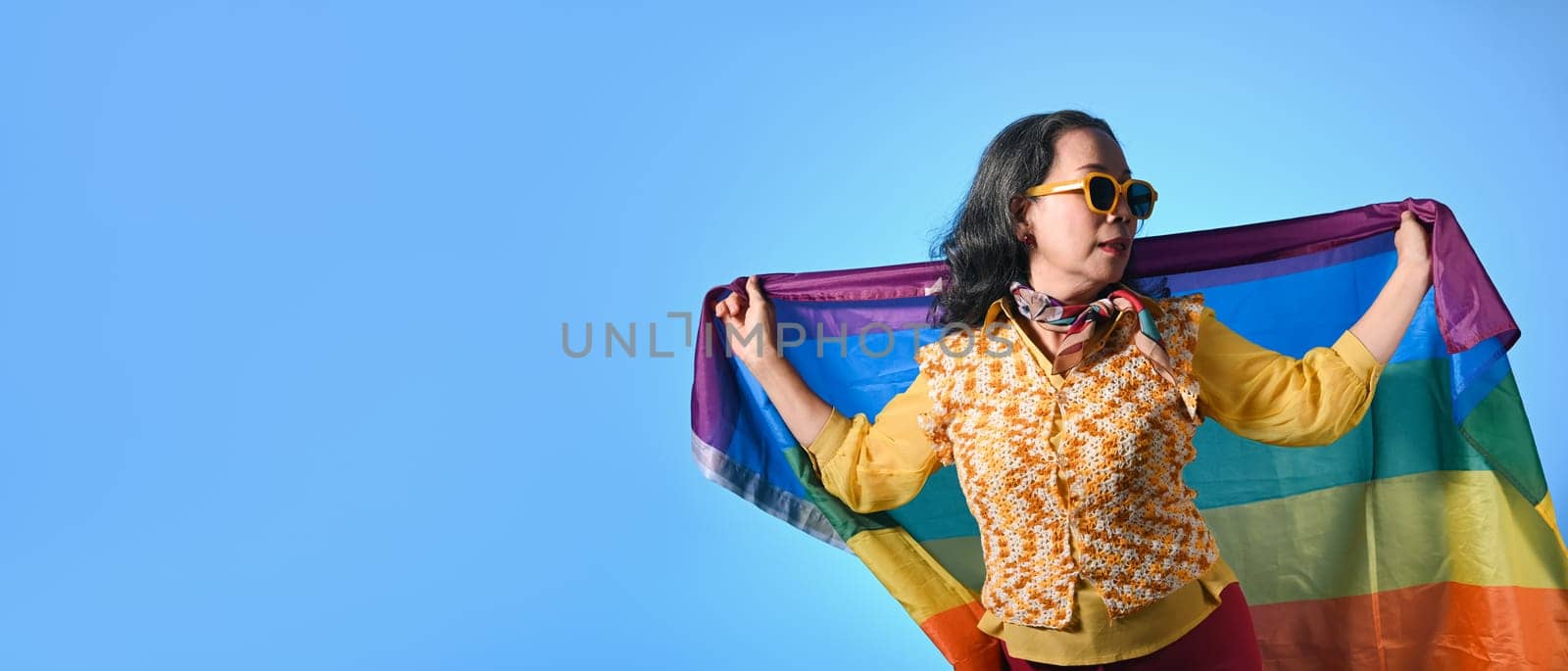 Beautiful mature woman waving rainbow flag on blue background. LGBT, human rights and equality concept.
