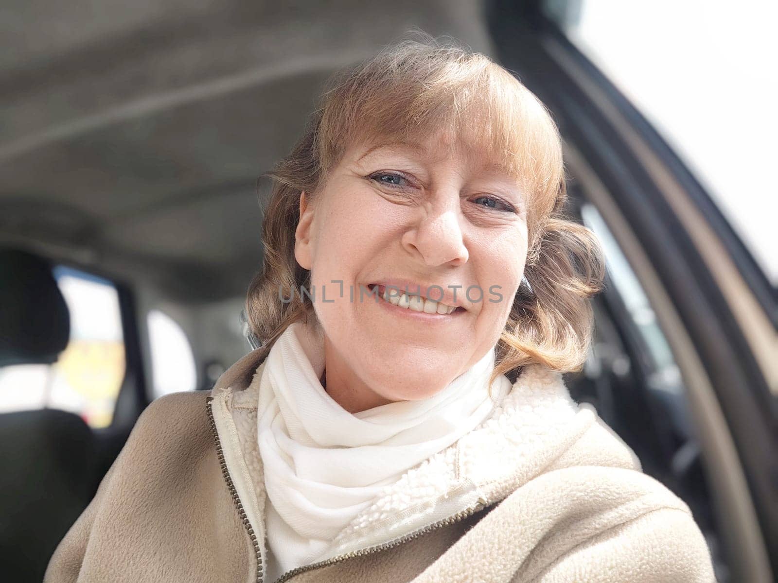 A middle-aged woman leans out of her car window, smiling as she takes selfie on a bright sunny day
