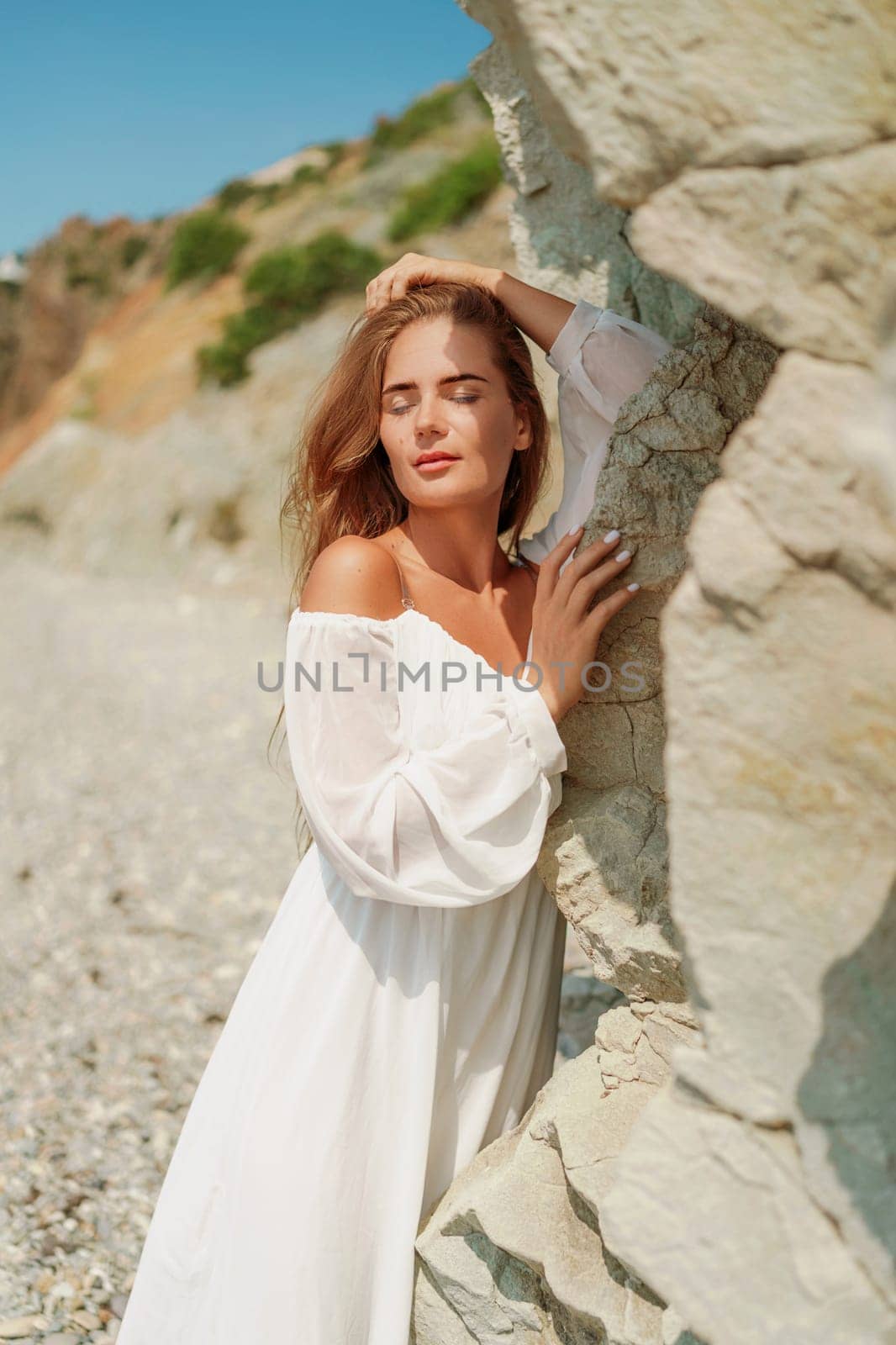 A woman in a white dress is standing on a rocky beach. Concept of serenity and tranquility, as the woman is enjoying the natural beauty of the beach