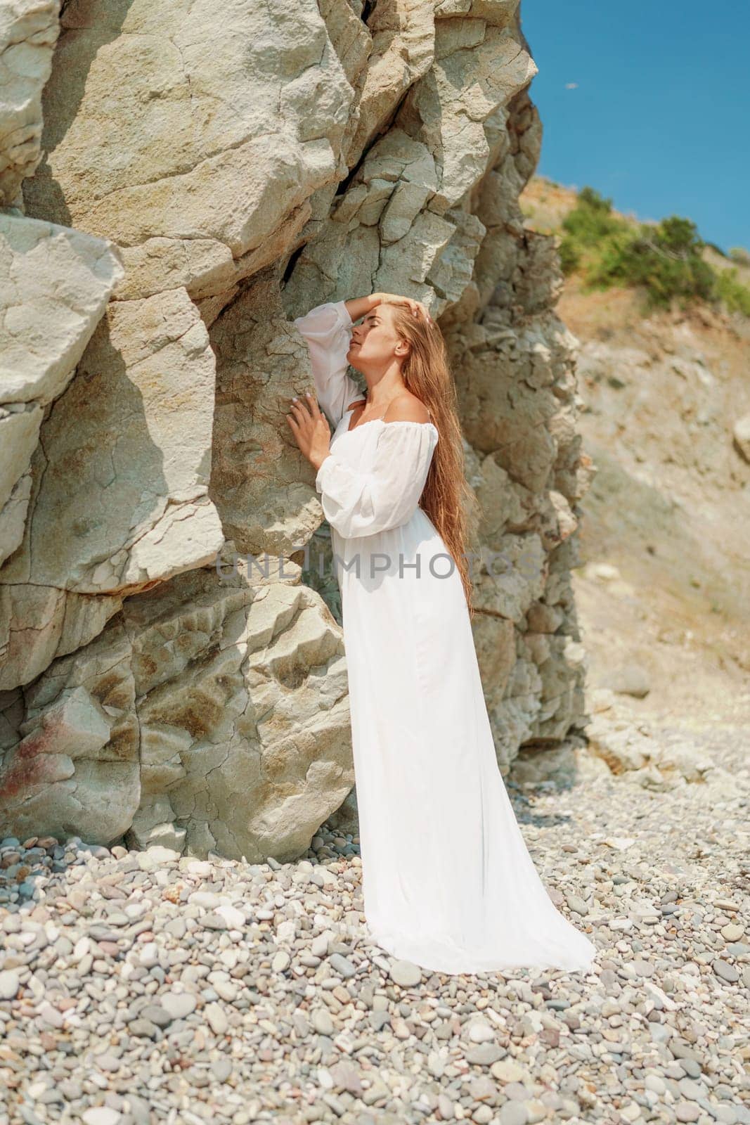 A woman in a white dress is standing on a rocky beach. Concept of serenity and tranquility, as the woman is enjoying the natural beauty of the beach. by Matiunina