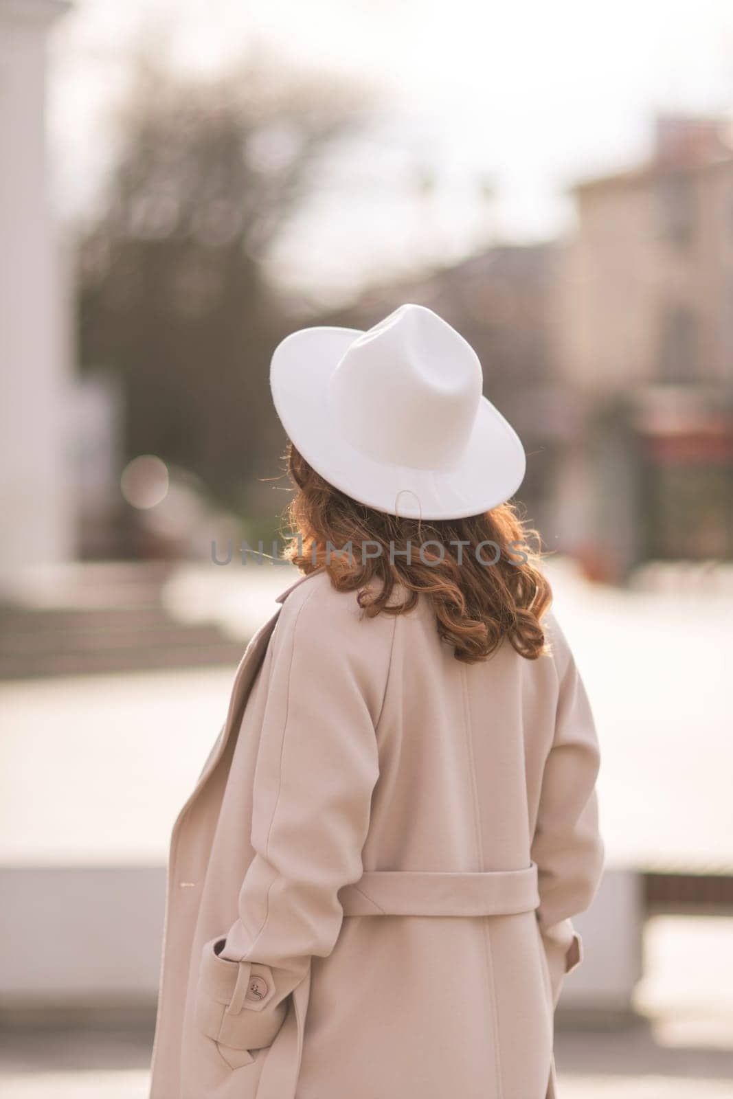 Back view of happy woman wearing hat and coat walking down street on sunny spring day. People, lifestyle, travel and vacations concept by Matiunina