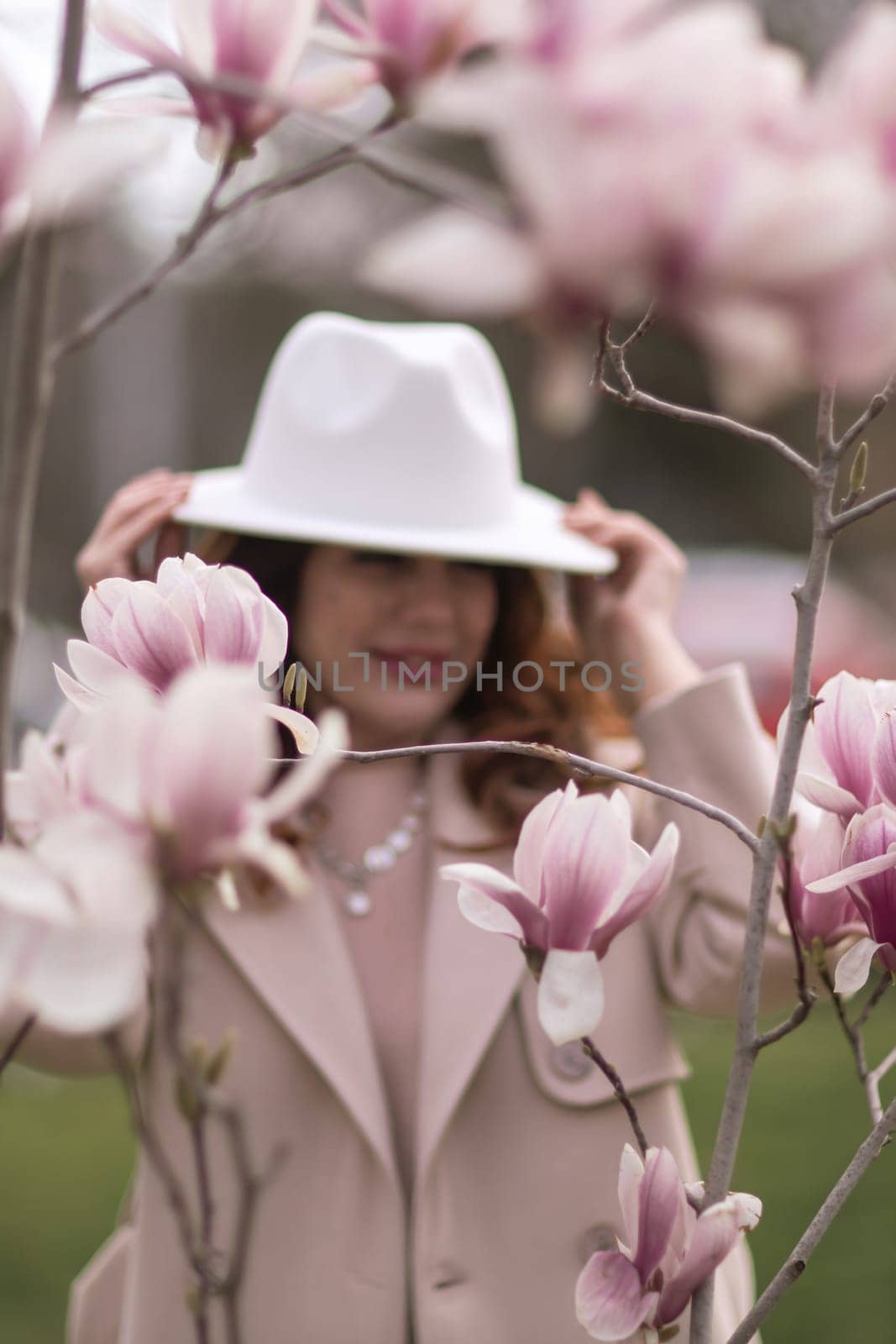 Woman magnolia flowers, surrounded by blossoming trees, hair down, white hat, wearing a light coat. Captured during spring, showcasing natural beauty and seasonal change