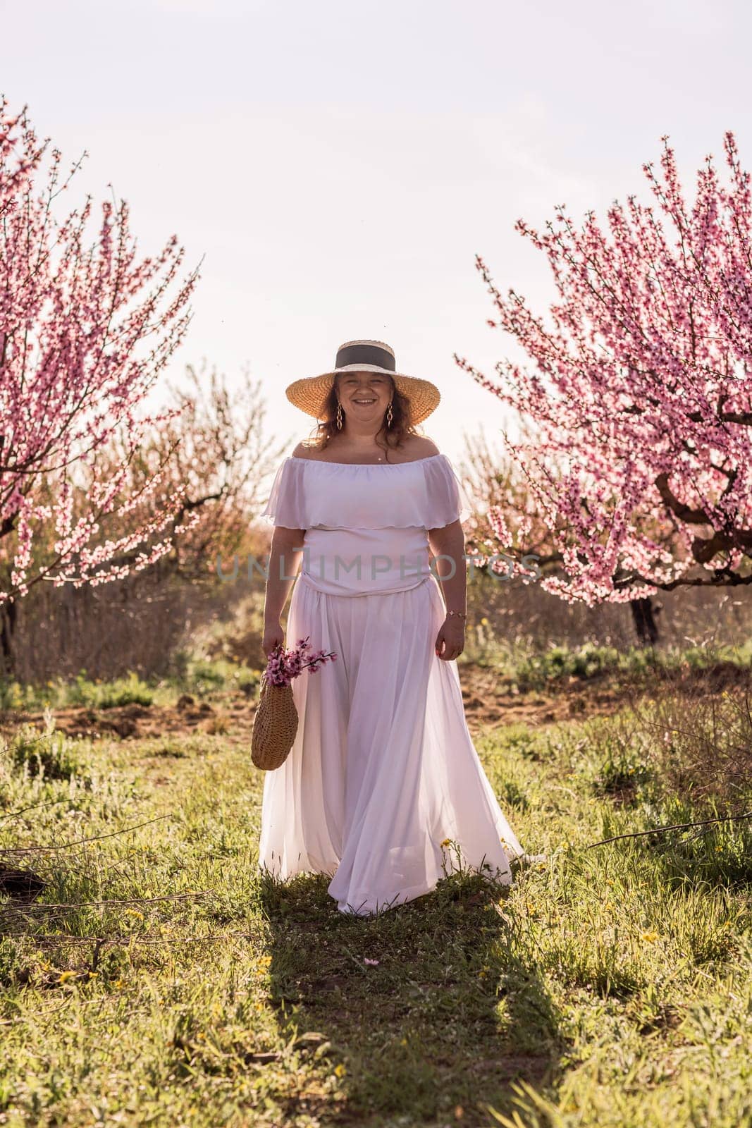 Woman blooming peach orchard. Against the backdrop of a picturesque peach orchard, a woman in a long white dress and hat enjoys a peaceful walk in the park, surrounded by the beauty of nature. by Matiunina