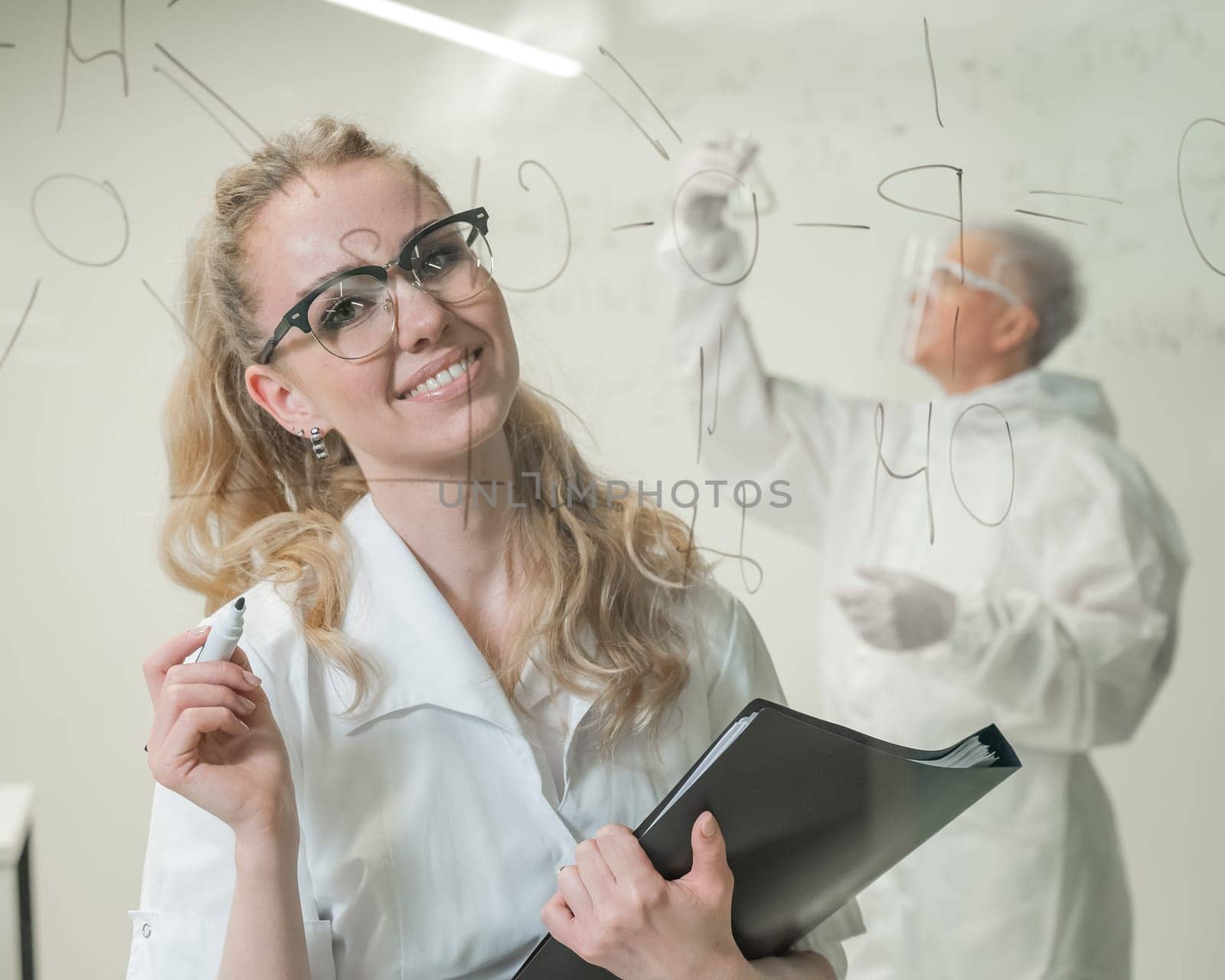 A woman chemist writes a formula on glass. An elderly Caucasian man in a protective suit is doing tests