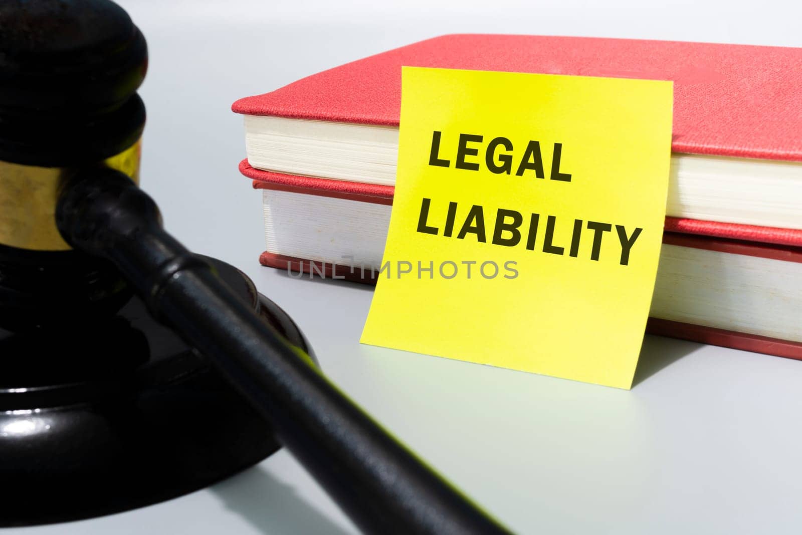 Judge's gavel with word legal liability written on yellow note and with red books on white background.