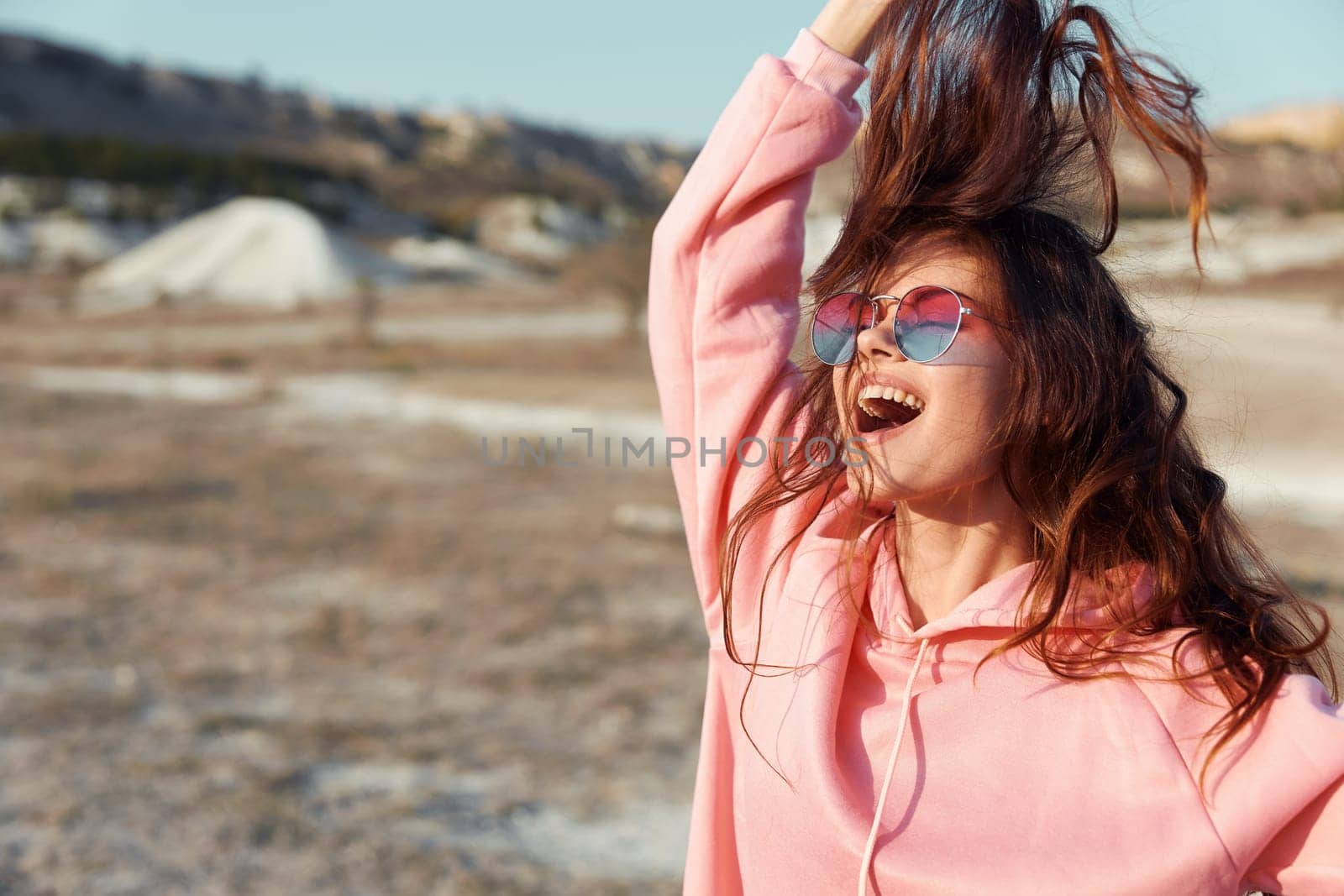 Stylish woman in pink sweatshirt and sunglasses standing in desert with wind blowing through her hair