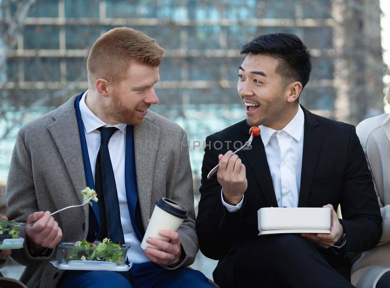 Front view of two multiethnic business colleagues men eating outdoors healthy home made salads.