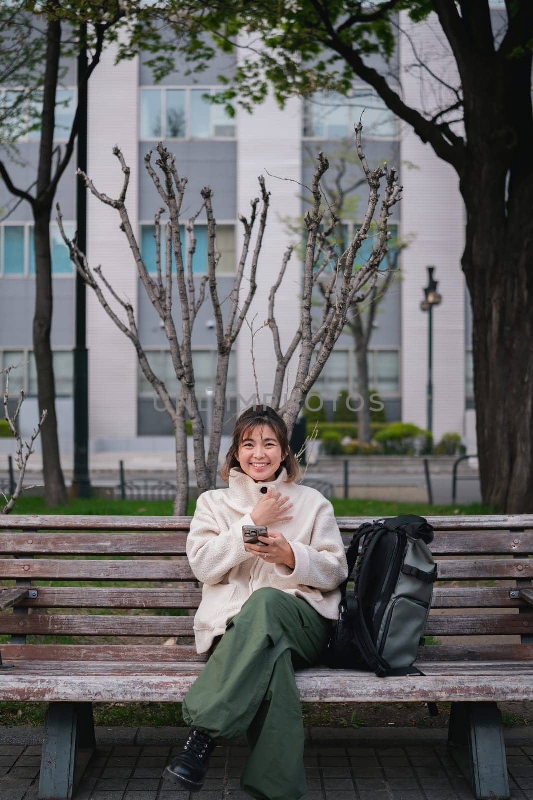 Woman sitting on a park bench smiling while using a smartphone with a backpack. Concept of outdoor relaxation and digital connectivity by wichayada