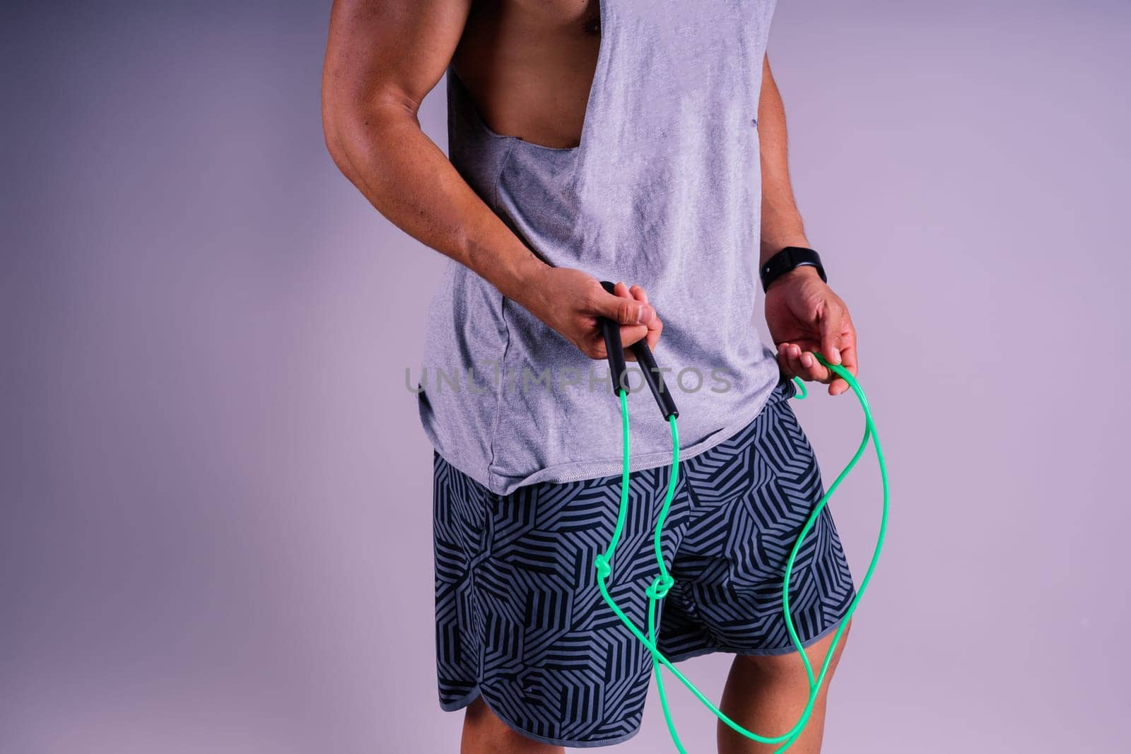 A man with a grey tank top is holding a green jump rope with his elbow bent and waist twisted, showing off his toned chest and wrist strength