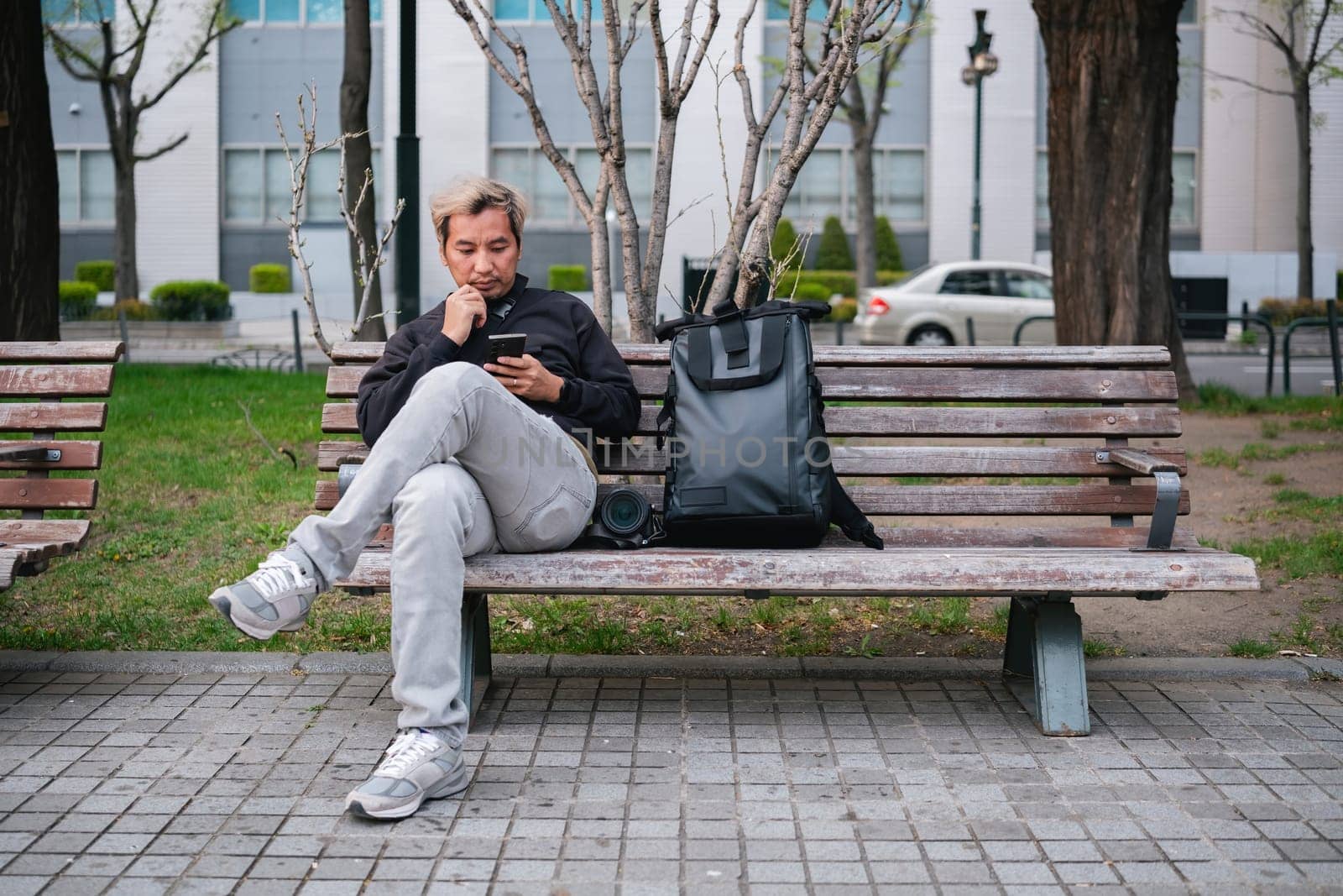 Man sitting on a park bench using smartphone with backpack and camera. Concept of urban relaxation and technology.