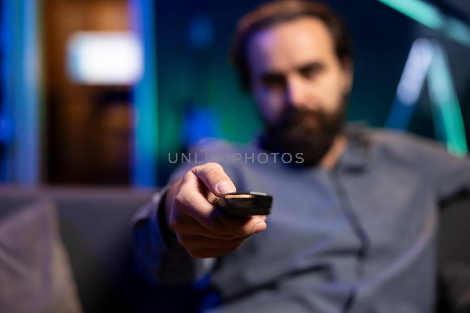 Close up shot of man in neon lit room browsing though movies available on streaming service, enjoying time off from work. Focus on remote control held by viewer in blurry background at home on couch