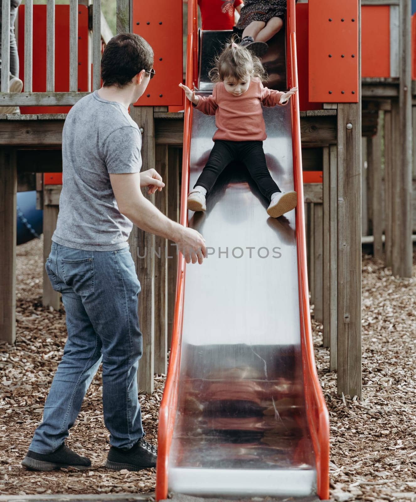 Portrait of one young Caucasian male father watching over his little daughter as she slides down a slide on a playground in a nature reserve on a sunny spring day in Belgium, close-up side view.