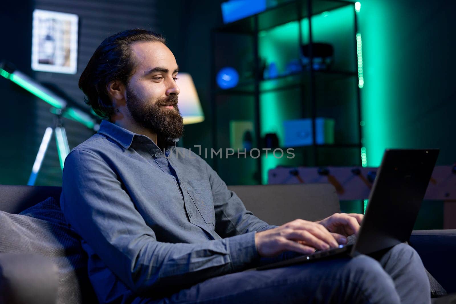 Smiling happy teleworker lying on couch, remotely performing tasks in personal home office. Cheerful relaxed freelancing man inputting data on laptop while working from home