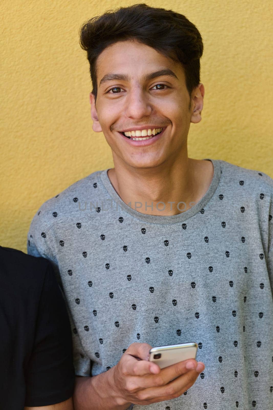 Middle Eastern teenager engrossed in his mobile phone while leaning against a vibrant yellow wall.