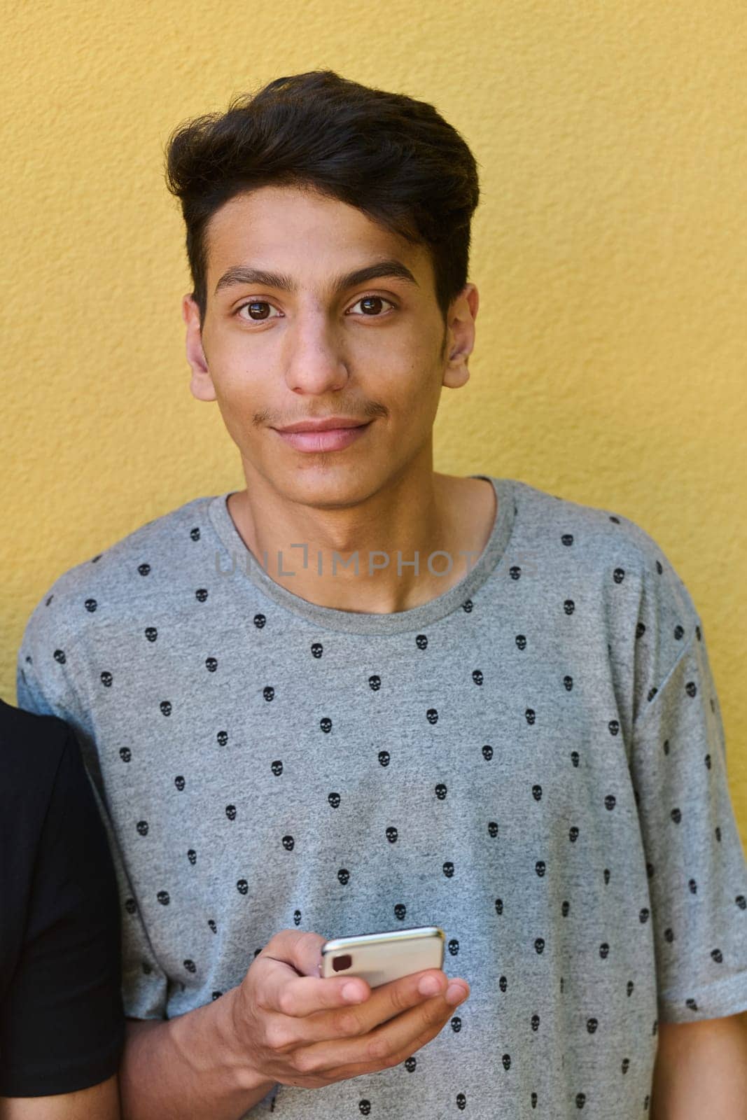 Middle Eastern teenager engrossed in his mobile phone while leaning against a vibrant yellow wall.