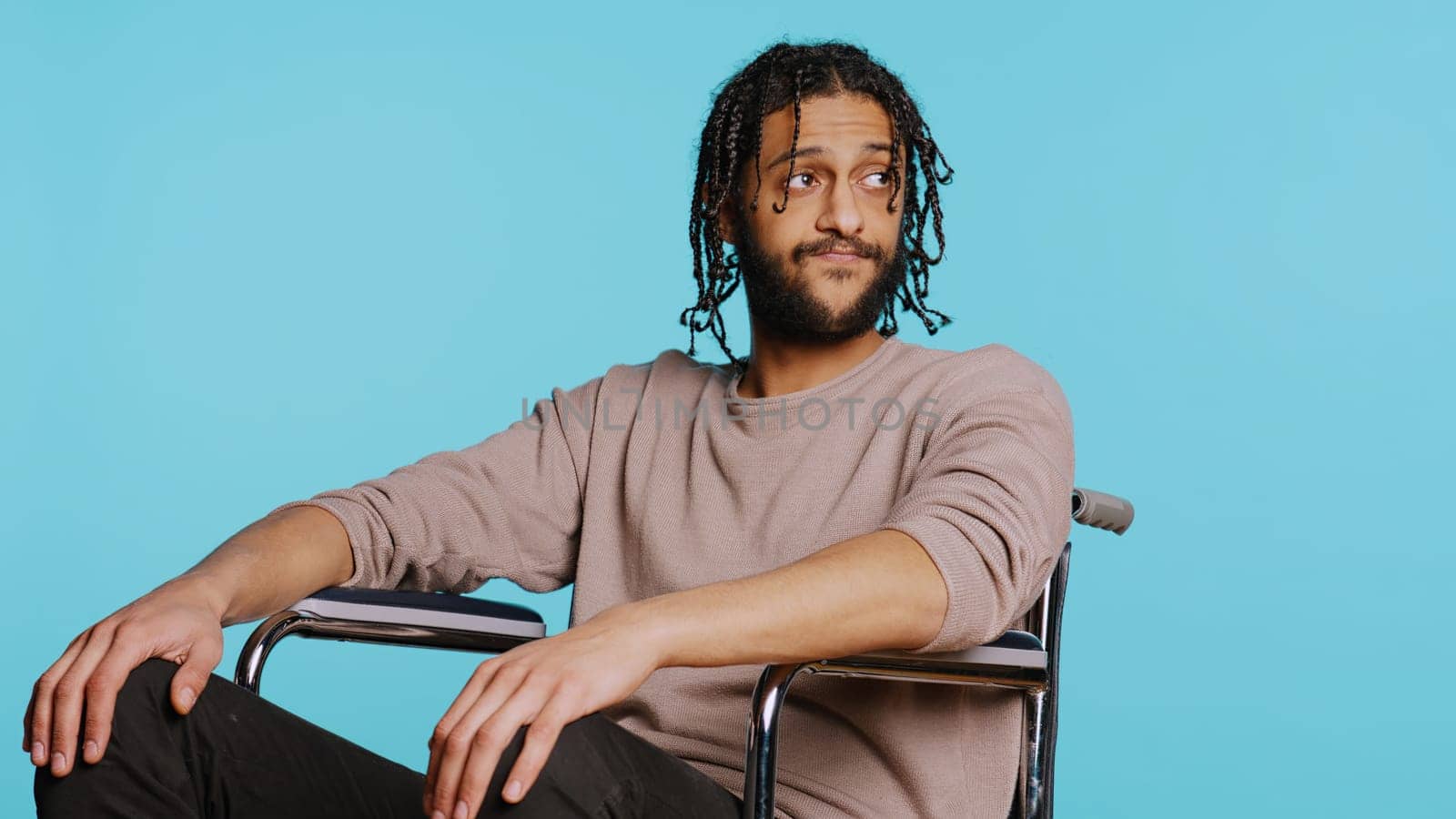 Bored Middle Eastern man with disability in wheelchair impatiently tapping fingers on knee, waiting. Annoyed person with paraplegia looking around, isolated over studio background, camera B