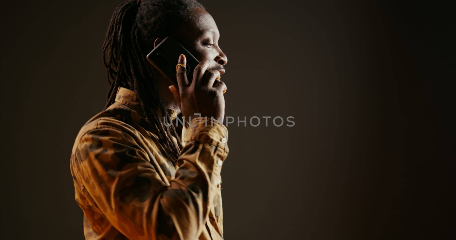 Cool guy with dreads answering phone call in studio, talking to friends or family on camera. African american trendy adult chatting remotely with people on telephone line. Handheld shot.