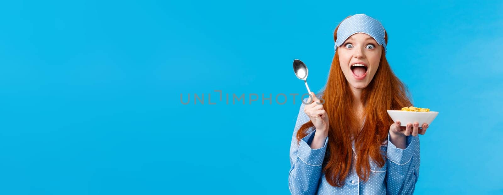 Excited, overwhelmed happy redhead caucasian woman eating cereals morning, holding spoon, smiling amused and amazed staring camera, wear sleep mask, pyjama, standing blue background.