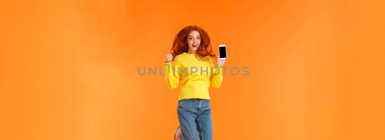 Advertising, technology and holidays concept. Vertical full-length shot cheerful gorgeous ginger girl, redhead woman jumping over orange background with smartphone, show thumb-up by Benzoix