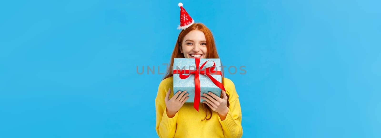 Girl cant wait give sibling her gift, smiling carry cute christmas wrapped present and wearing fancy santa hat, laughing, sharing positive emotions, attend winter holidays party, blue background.