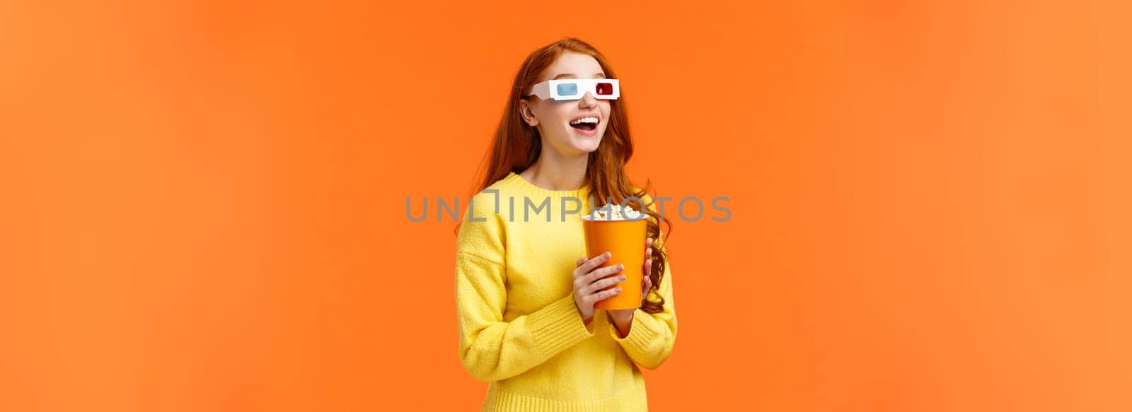 Girl eating popcorn, smiling amused as staring at large screen watching movie at cinema, open mouth thrilled, wear 3d glasses on fantasy film theatre, standing orange background.
