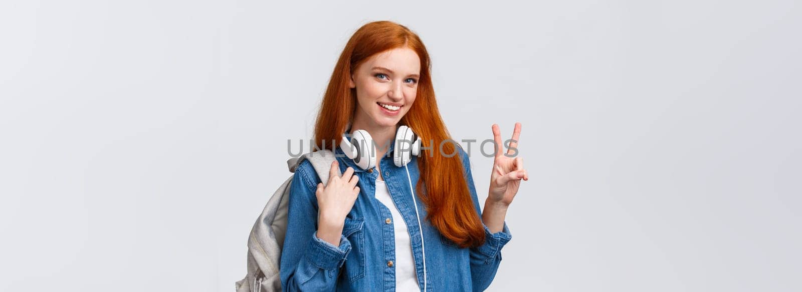 Lovely charismatic caucasian redhead girl with backpack, headphones over neck, showing peace sign and smiling delighted and joyful camera, standing white background, heading part-time job.