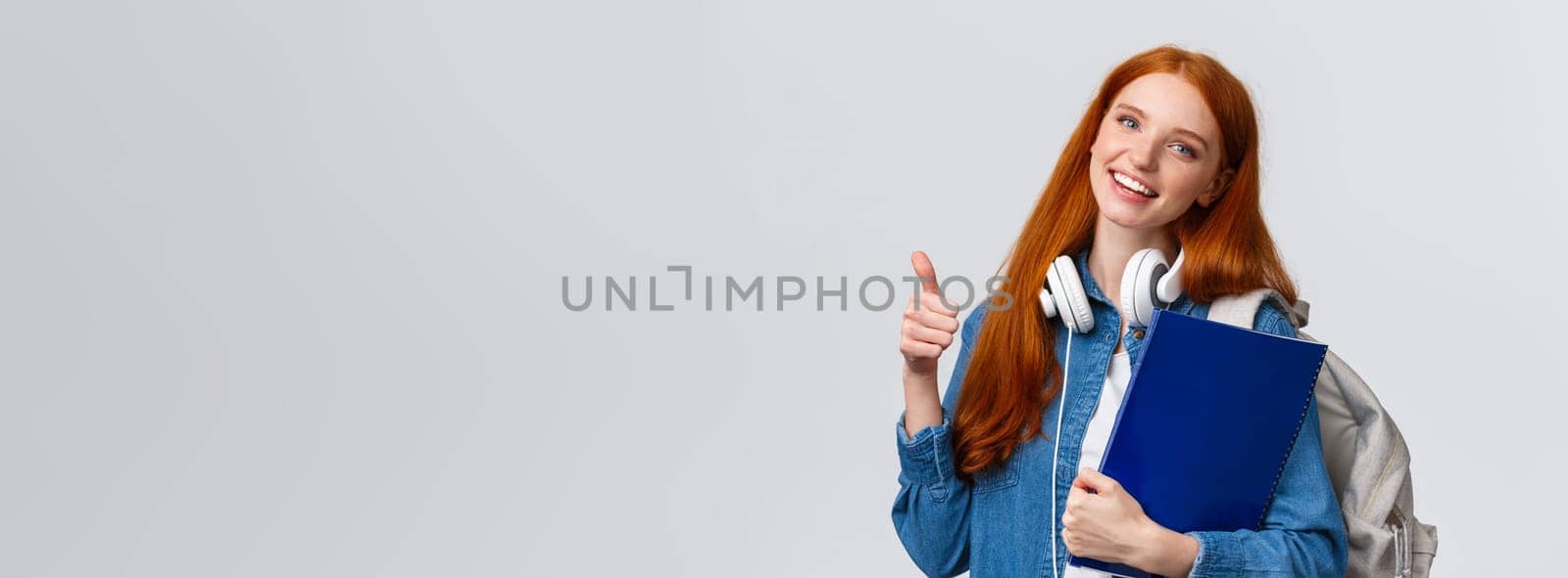 Education, teenagers and student lifestyle concept. Cheerful redhead lovely girl with backpack and folder, bring her portfolio to art, design class, showing thumb-up in approval, like gesture.