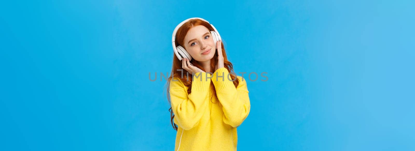 Waist-up shot tender and cute, lovely redhread woman in yellow sweater, tilt head, wear headphones, touching earphones as press to ears, listen music, smiling camera delighted, blue background.